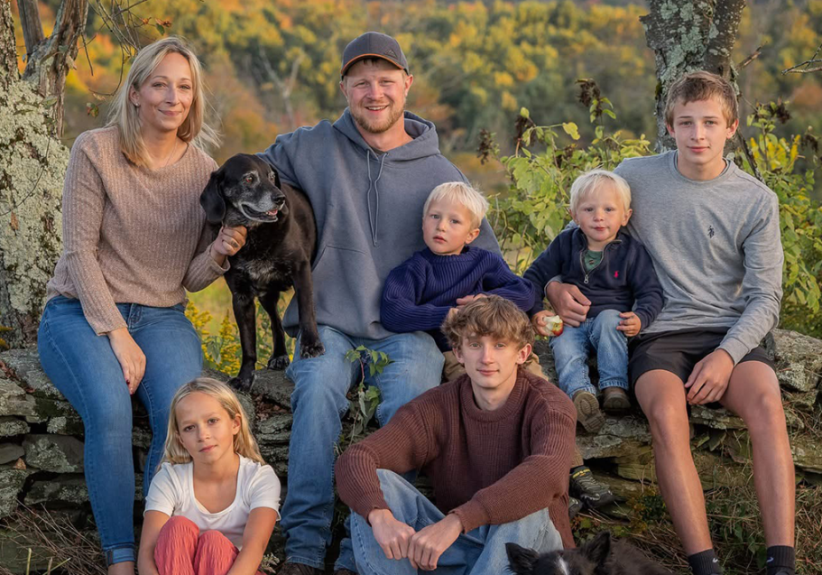 Family photo featuring parents, five children and a black dog sitting in the woods