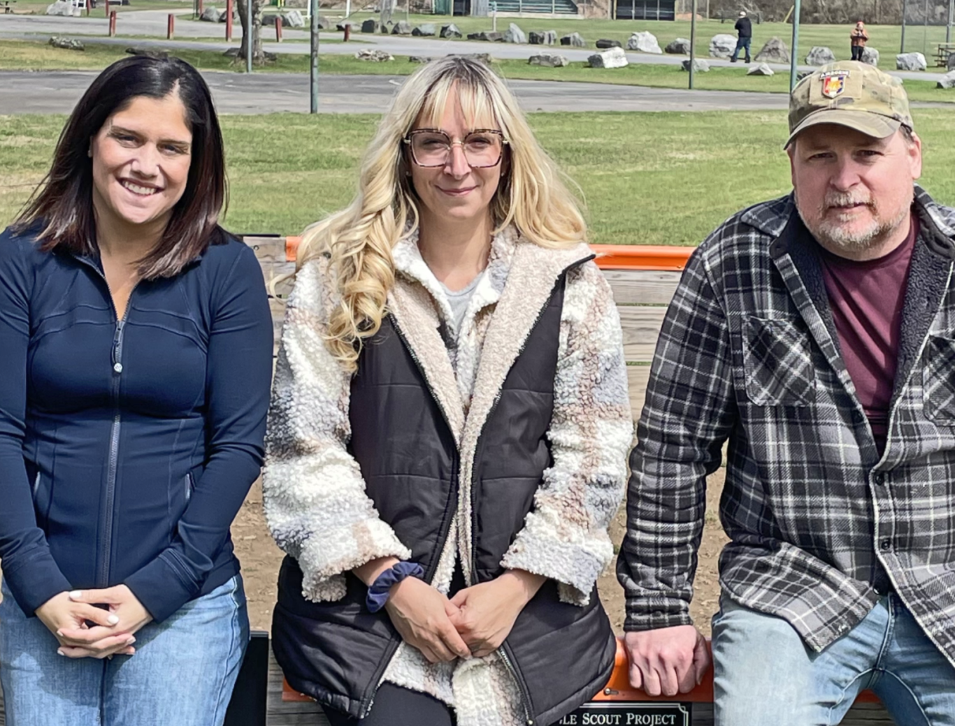 Two women and one man sit on a fence in a community park